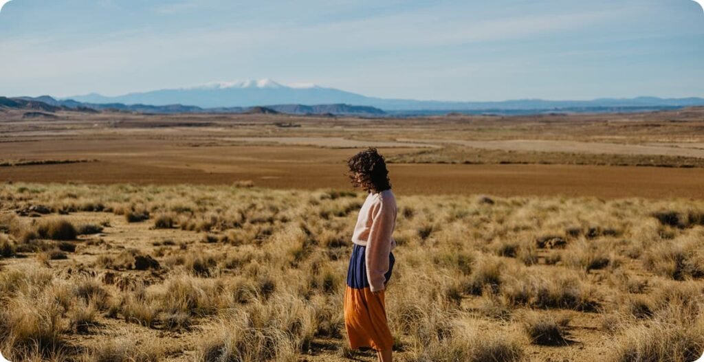 Person standing in wide semi-arid plain with dry grass, distant hills, and snow-capped mountains under clear sky.