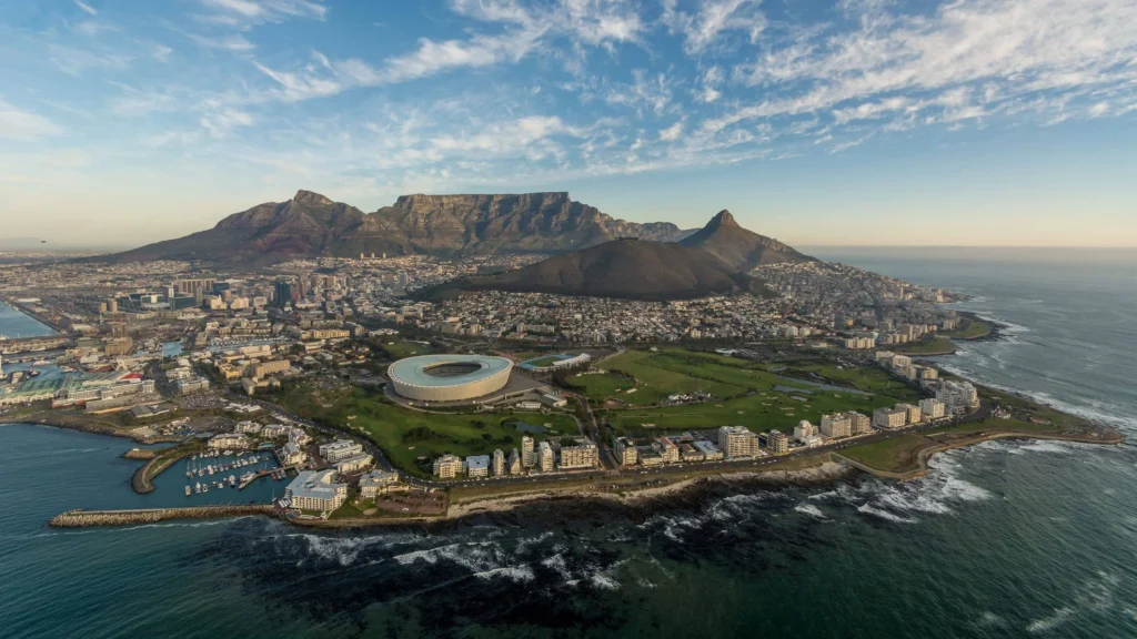 Aerial view of Cape Town showing circular stadium near coastline with Table Mountain and Lion’s Head in background.