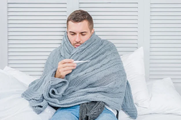 Person wrapped in a gray blanket sitting on a bed and checking a thermometer, suggesting fever or illness.