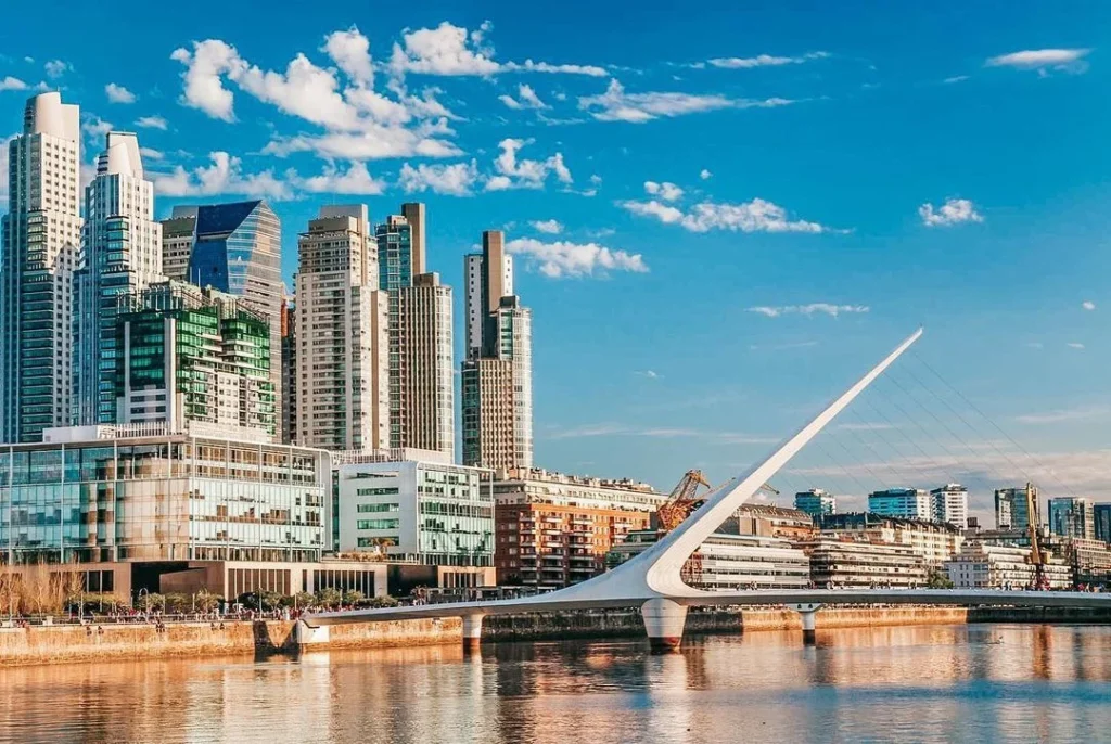 Modern urban skyline of Buenos Aires with tall skyscrapers and distinctive white Puente de la Mujer bridge reflected in waterfront.