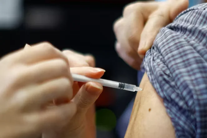 A close-up of a healthcare provider's hands holding a syringe, about to administer a vaccine into the upper arm of a patient wearing a plaid shirt.