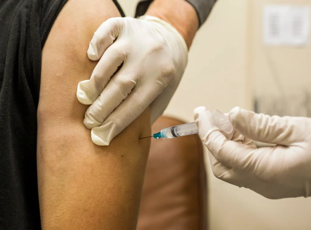 Close-up of a syringe injecting into a person’s upper arm in a clinical setting.