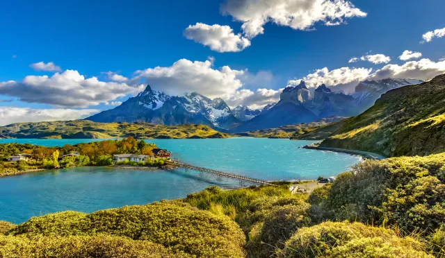 Turquoise lake surrounded by lush green hills and snow-capped mountains, with wooden bridge leading to lodge under bright blue sky.