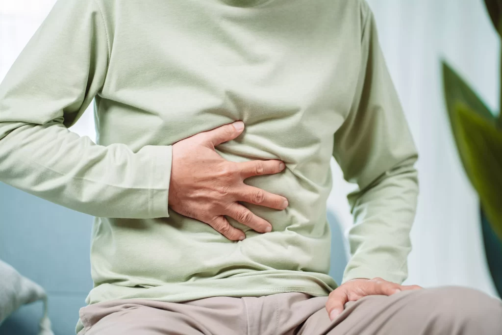 Person in a green shirt sitting and pressing their upper abdomen, showing signs of abdominal discomfort.