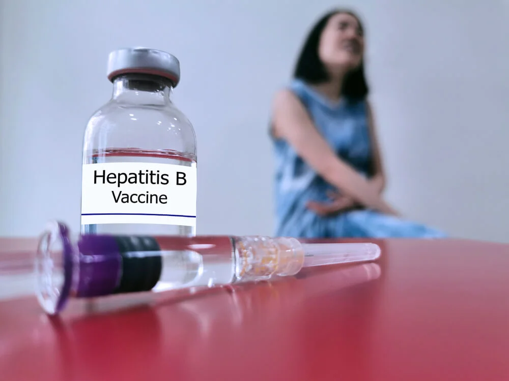 Vial labeled “Hepatitis B Vaccine” beside syringe on red surface with blurred person in background.