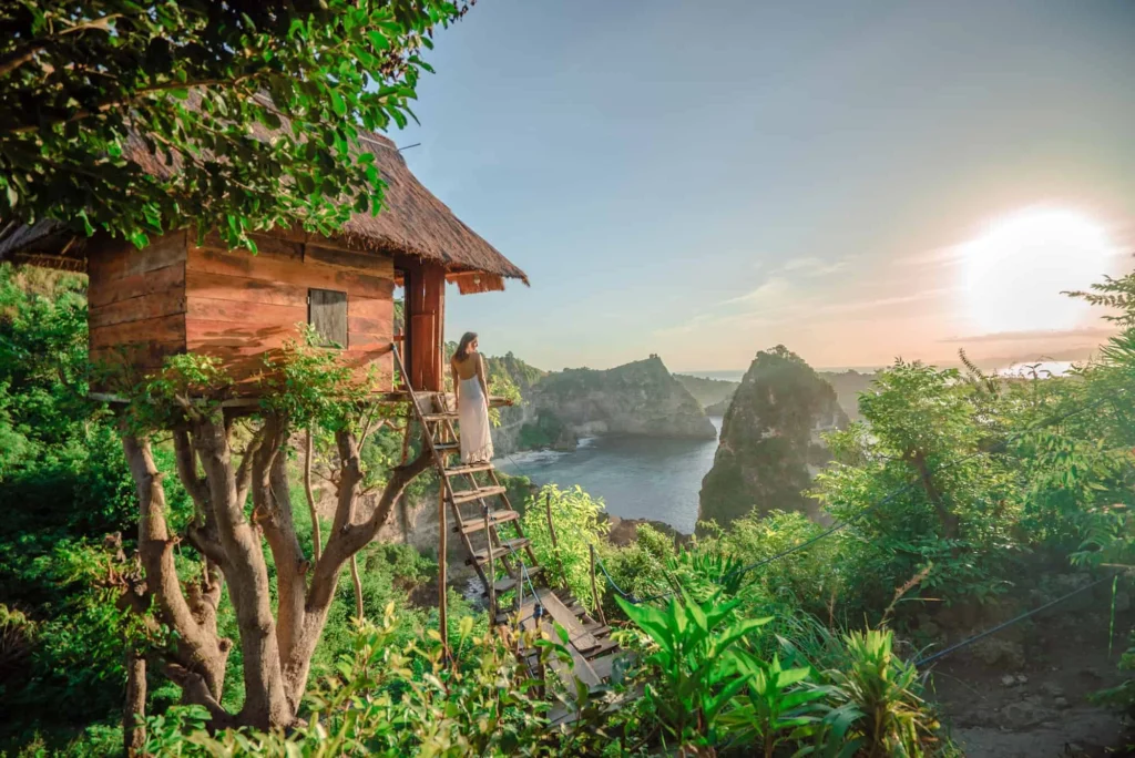 Scenic coastal treehouse with thatched roof perched among greenery, person in white dress on ladder overlooking ocean cliffs and islands at sunrise.