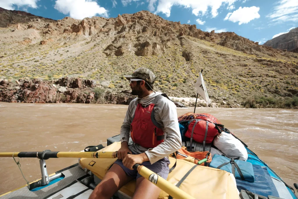 Person in red life vest rowing gear-loaded raft on muddy river through rocky, mountainous terrain under partly cloudy sky.