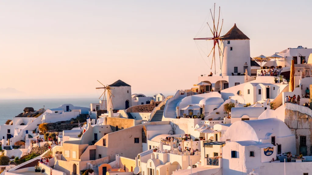 Picturesque coastal town in Santorini, Greece with whitewashed buildings, blue domes, windmills, and golden sunset over the sea.