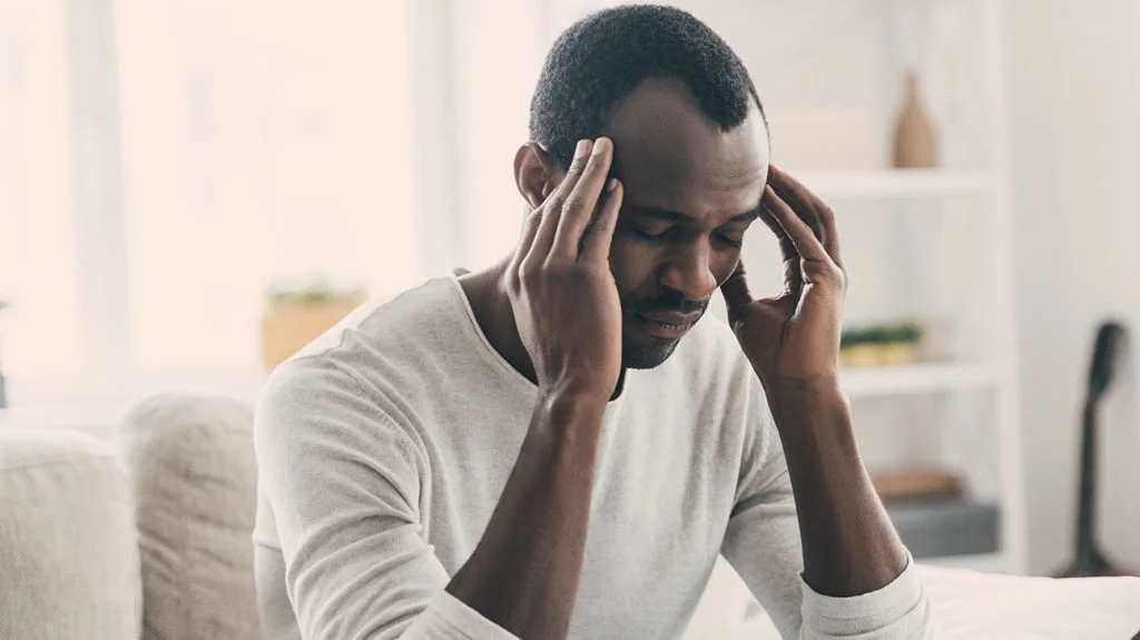 Person sitting indoors with hands on temples, suggesting headache or stress.