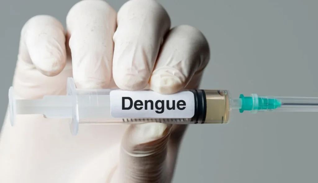A gloved hand holding a syringe labeled "Dengue" containing a yellowish liquid.