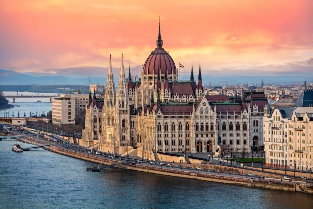 Hungarian Parliament Building along Danube River at sunset with glowing sky and cityscape.