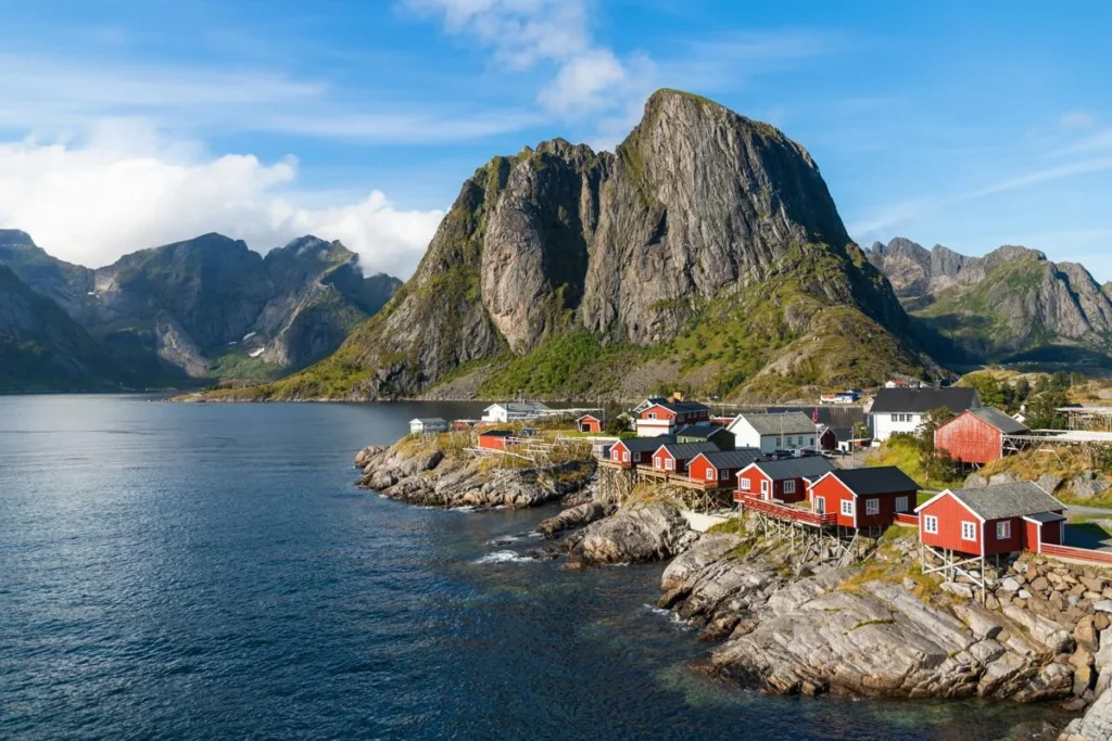 Scenic coastal village with red and white wooden houses on stilts along rocky shoreline, backed by steep mountains and calm water.