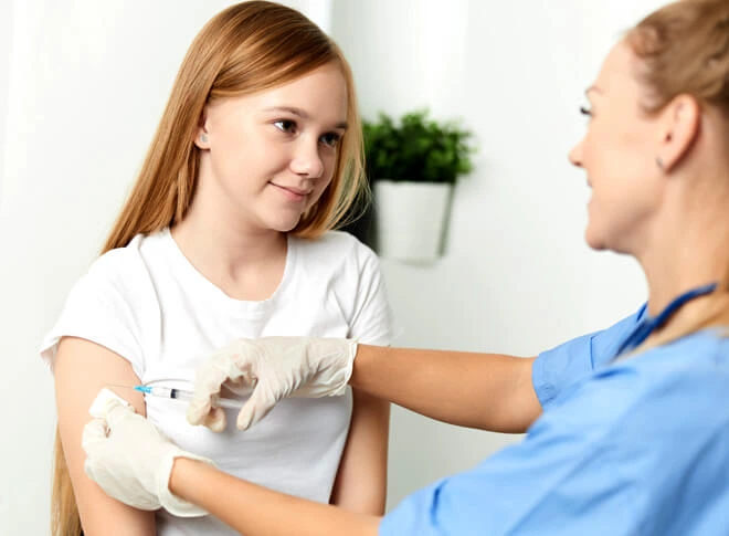 A young girl smiling while receiving a vaccination in her arm from a nurse in blue scrubs.