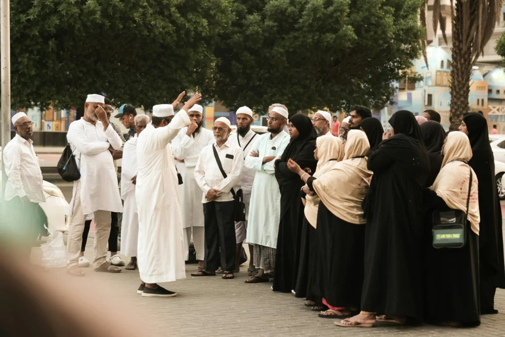 Group of people in traditional attire gathered outdoors, with one person addressing the group near trees and buildings.
