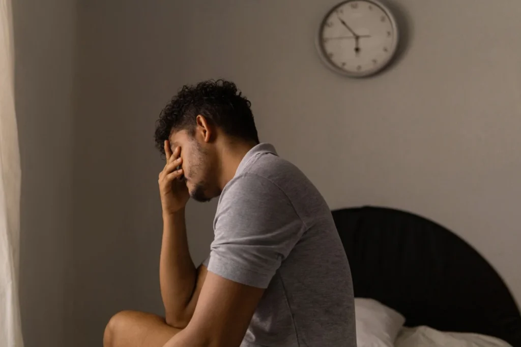 Person sitting on bed in dimly lit room with hand covering face near window and clock.