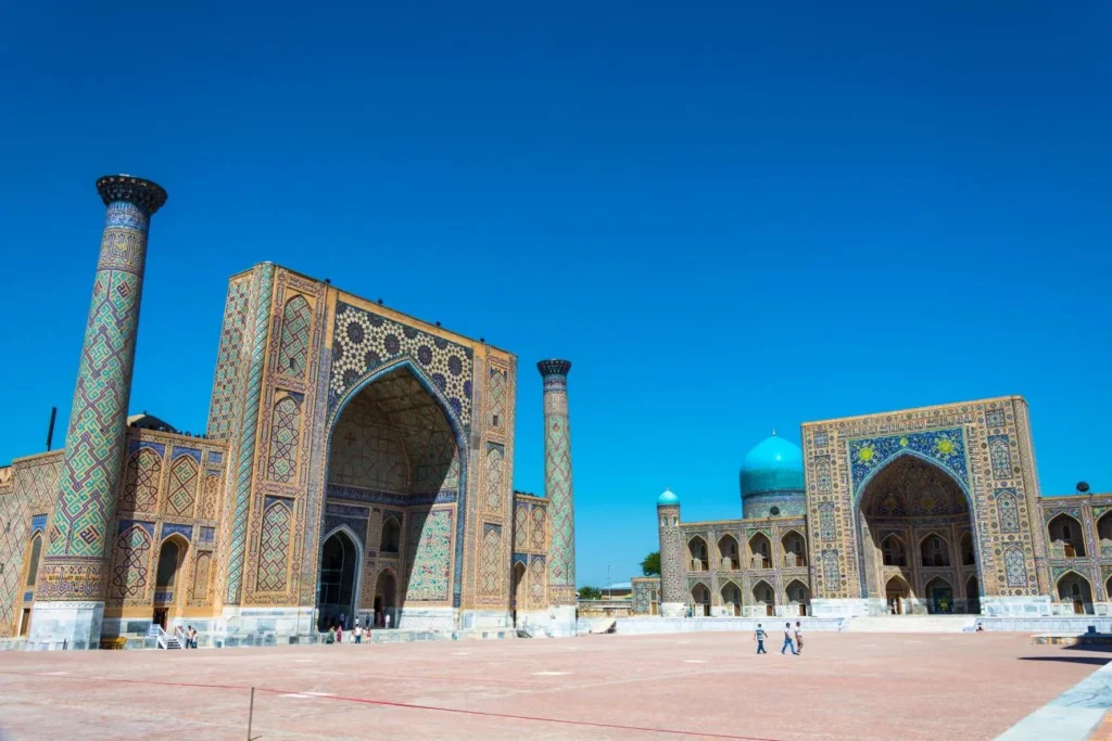 Grand Islamic architectural complex with intricate tilework, tall minarets, turquoise dome, and paved courtyard under clear blue sky.
