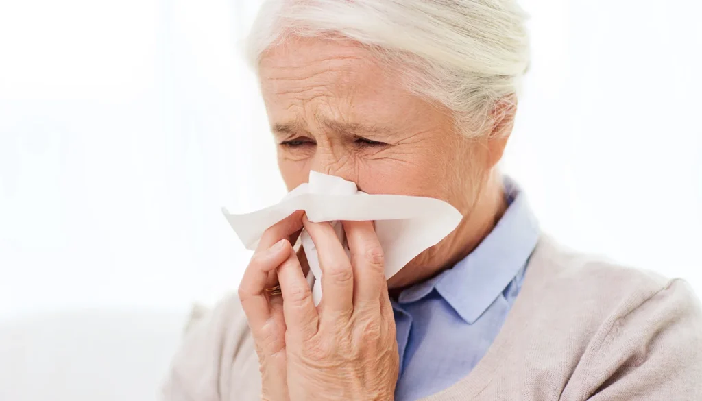 Older adult in a beige sweater holding a tissue to the nose, appearing to sneeze or wipe.