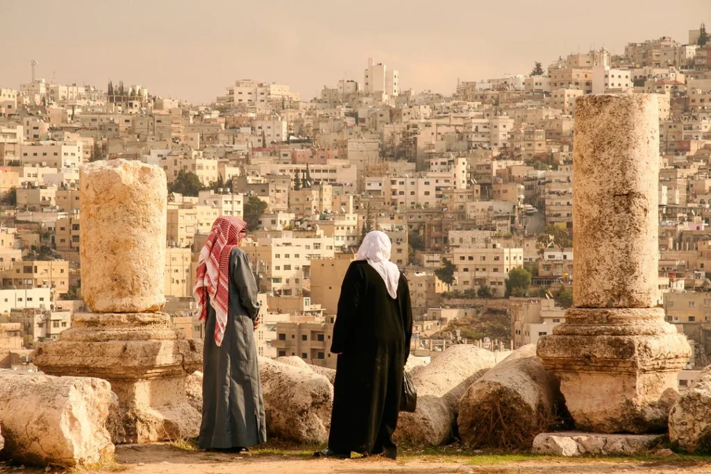 Two individuals in traditional Middle Eastern attire standing among ancient stone ruins with panoramic view of hillside city of beige buildings.