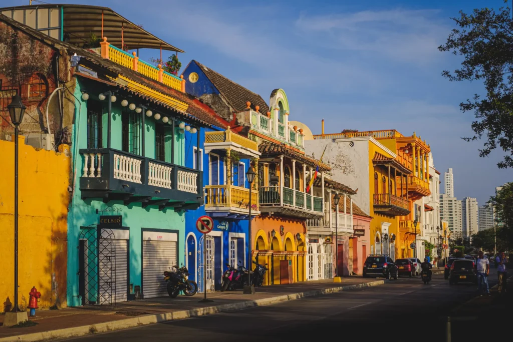 Vibrant street lined with colorful colonial-style buildings featuring ornate balconies, shutters, and tiled roofs, with parked motorcycles and pedestrians under warm sunlight.