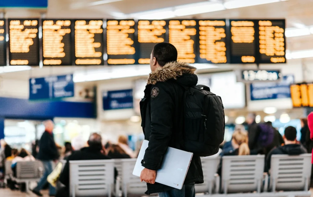 Traveler in airport terminal wearing backpack and fur-lined jacket, holding laptop while looking up at large electronic departure board.
