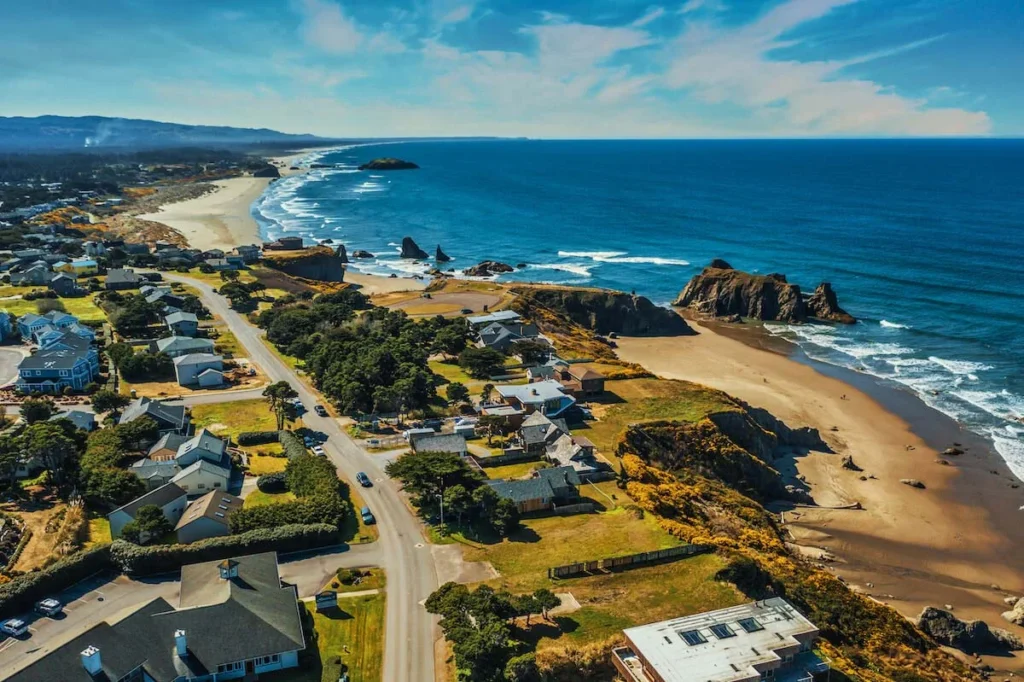 Aerial view of coastal town with sandy beach, rocky shoreline, residential houses, and ocean waves stretching to horizon.