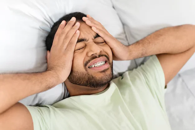 Person lying on a bed with hands on the head and eyes closed, showing signs of headache or stress.