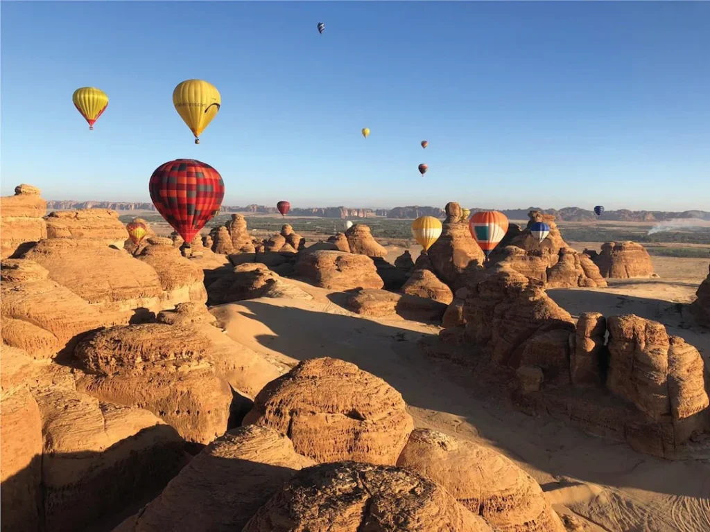 Desert landscape with eroded sandstone formations and colorful hot air balloons floating in clear blue sky.
