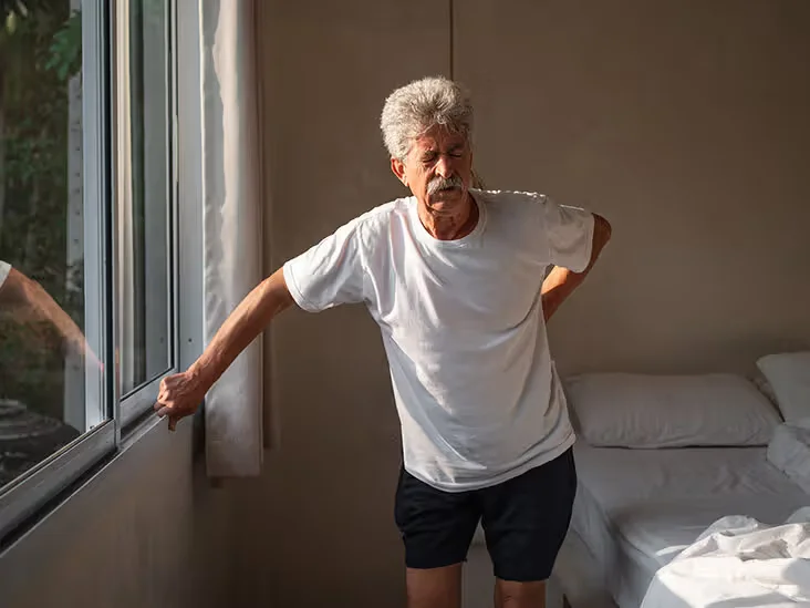 Older adult standing by bed holding lower back, suggesting discomfort or stiffness.