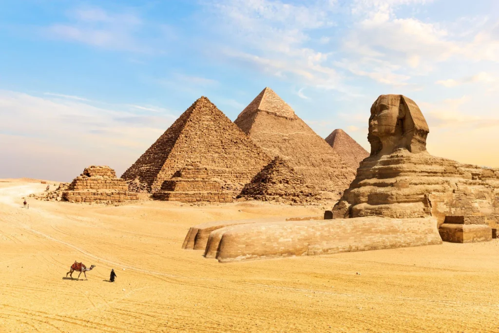 Desert landscape with Great Sphinx of Giza in foreground and three pyramids rising behind under clear blue sky.