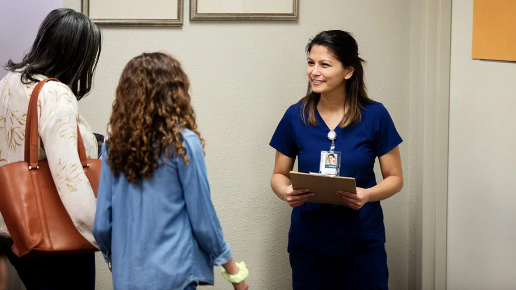 A smiling nurse in blue scrubs holds a clipboard while greeting a woman and a young girl in a clinic.