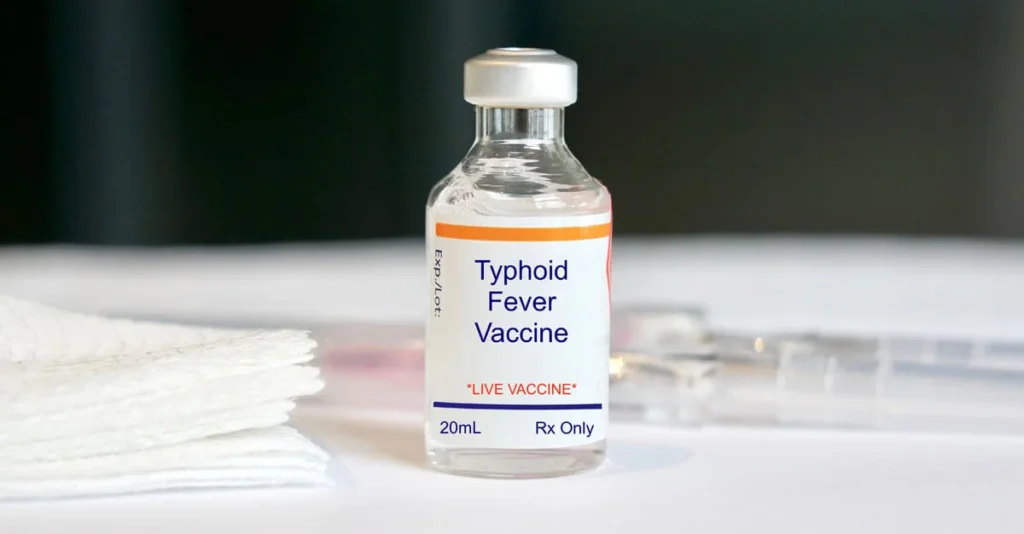 Small glass vial labeled “Typhoid Fever Vaccine” placed on a white surface with gauze and a syringe.