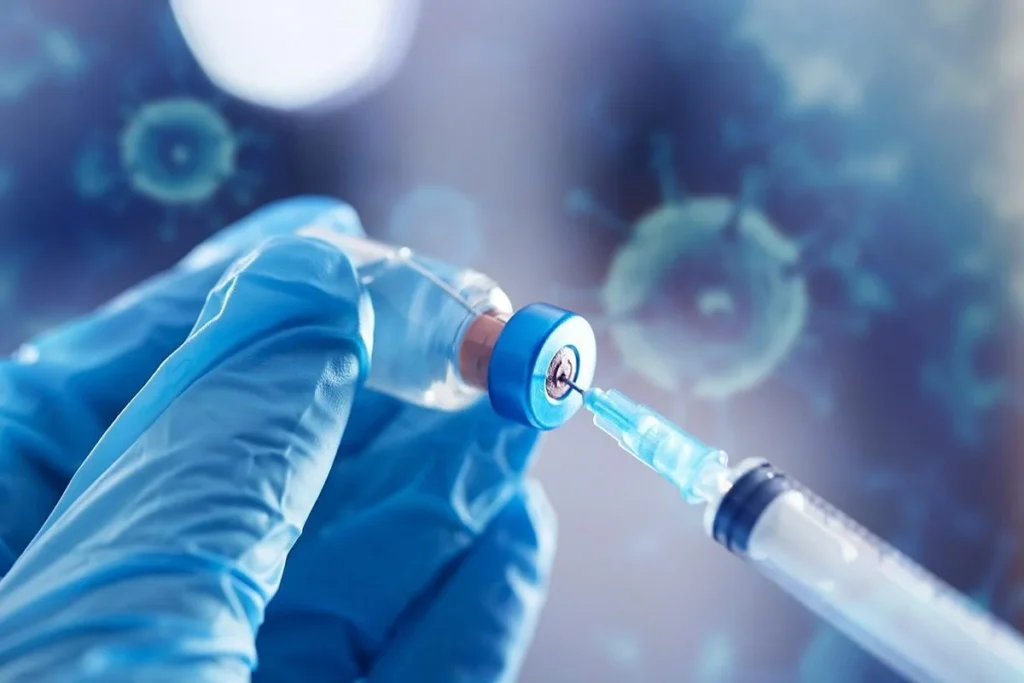 Gloved hand inserting syringe needle into a vial with virus illustrations in the background.