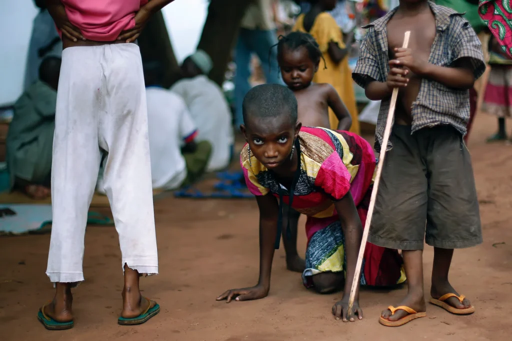A young girl in a colorful dress crawls on red-brown earth, looking intensely at the camera. She is surrounded by other children in a crowded, shaded outdoor area.
